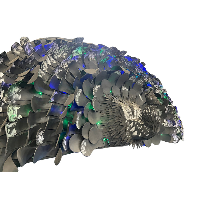 A close-up of a metallic, scale-like decorative object with layered dark gray and silver leaf or petal shapes, accented by glowing blue and green lights, shown against a plain white background.